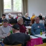 Menschen sitzen an Tischen in einer Halle, essen und unterhalten sich.