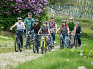 Gruppe aus Erwachsenen und Kindern auf Fahrrädern bei einer geführten Radtour durch eine blühende Frühlingslandschaft.