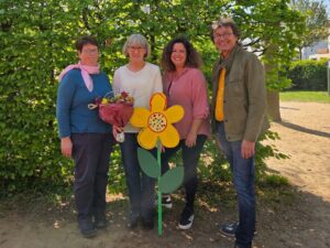 Vier Personen stehen lachend vor einer grünen Hecke. Eine Frau hält einen Blumenstrauß, daneben eine große, bunte Holzblume mit Botschaften.