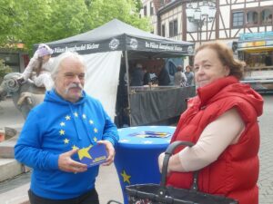 Mann im blauen EU-Hoodie mit einer Frau am Marktstand, hält eine Europatag-Postkarte.