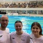Drei Personen in weißen Vereins-Shirts stehen lächelnd vor einem Schwimmbecken in einer großen Schwimmhalle mit Zuschauertribüne.