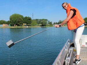 Eine Frau mit Schwimmweste nimmt mit einer langen Stange eine Wasserprobe aus einem ruhigen Badesee vom Boot aus.