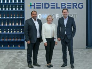 Drei Personen in Businesskleidung stehen vor einer Wand mit dem Schriftzug „Heidelberg – Home of Print“; links ein Regal mit Getränkeflaschen.