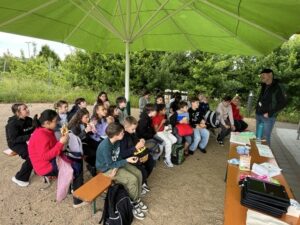 Schülergruppe unter einem grünen Schirm bei Vortrag über Nachhaltigkeit, einige Kinder essen Brezeln