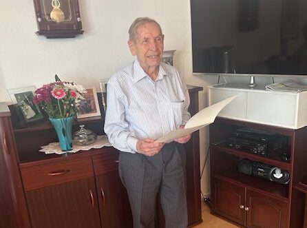 Ein älterer Herr steht in einem Wohnzimmer und hält eine Urkunde in den Händen. Er trägt ein Hemd und lächelt. Im Hintergrund sind Blumen, ein Fernseher und ein Schrank zu sehen.