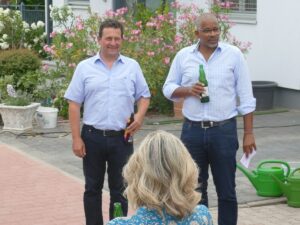 Zwei Männer mit Getränken in der Hand sprechen vor einer kleinen Gruppe auf einem Straßenfest.