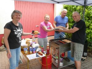 Vier Erwachsene an einem Grillstand reichen sich Bratwurstbrötchen, mit Getränken und Ketchup auf einem Tisch daneben, freundliche Stimmung im Freien.