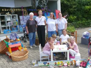 Gruppenfoto von Erwachsenen und Kindern vor einem Bastelladen mit Flohmarktspielzeug und Kindertisch – mehrere Generationen gemeinsam beim Verkauf.