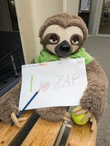 Plüschfaultier Fauli hält ein Blatt Malpapier mit der Aufschrift „I love ZAP“ und einem Buntstift auf einer Holzbank.